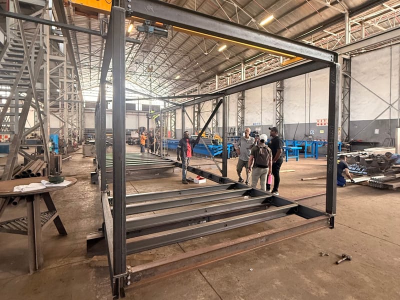 Container-sized steel frame being fabricated in the CSI workshop, with a crane overhead and team members inspecting the structure